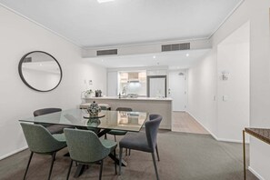 A modern kitchen with white cabinets, a central island, and a dining area with a glass table and chairs. - Spacious 3BR | Pool, Gym, Tennis & Parking (Brisbane)