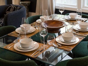 Mesa de jantar posta com pratos, tigelas e talheres brancos, rodeada por cadeiras estofadas verdes.