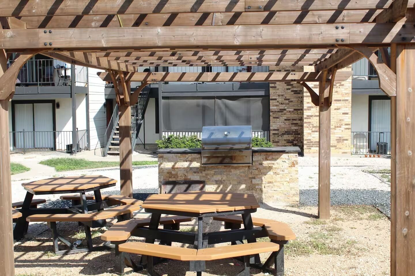 An outdoor patio with wooden tables and benches, a barbecue grill, and a pergola structure.