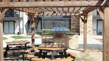 An outdoor patio with wooden tables and benches, a barbecue grill, and a pergola structure.