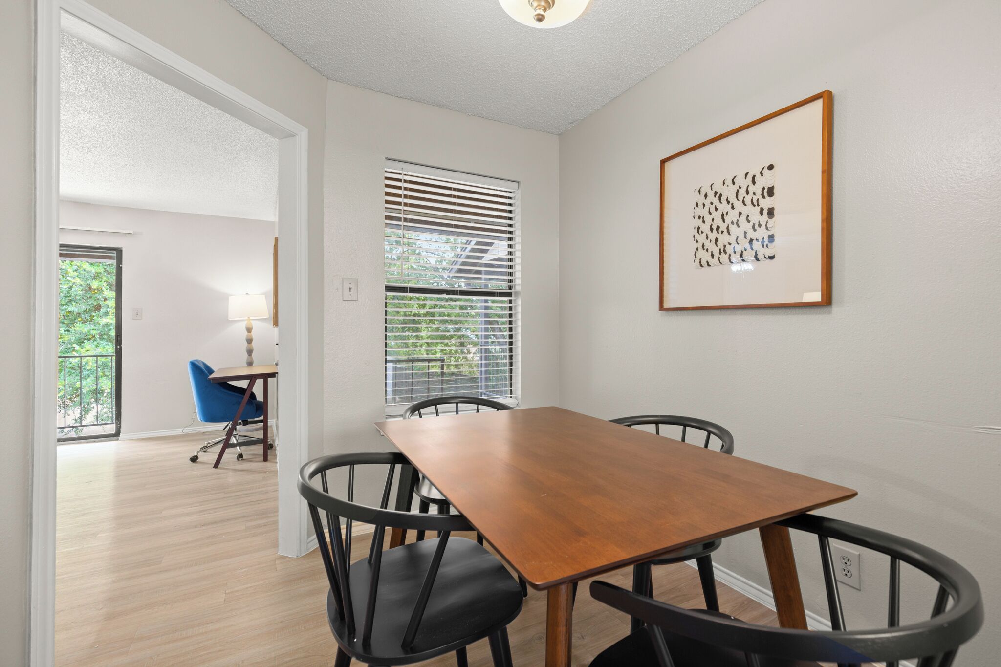 A dining area with a wooden table and four chairs, a window with blinds, and a framed artwork on the wall.