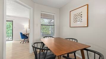 A dining area with a wooden table and four chairs, a window with blinds, and a framed artwork on the wall.