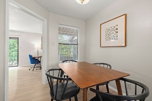 A dining area with a wooden table and four chairs, a window with blinds, and a framed artwork on the wall.