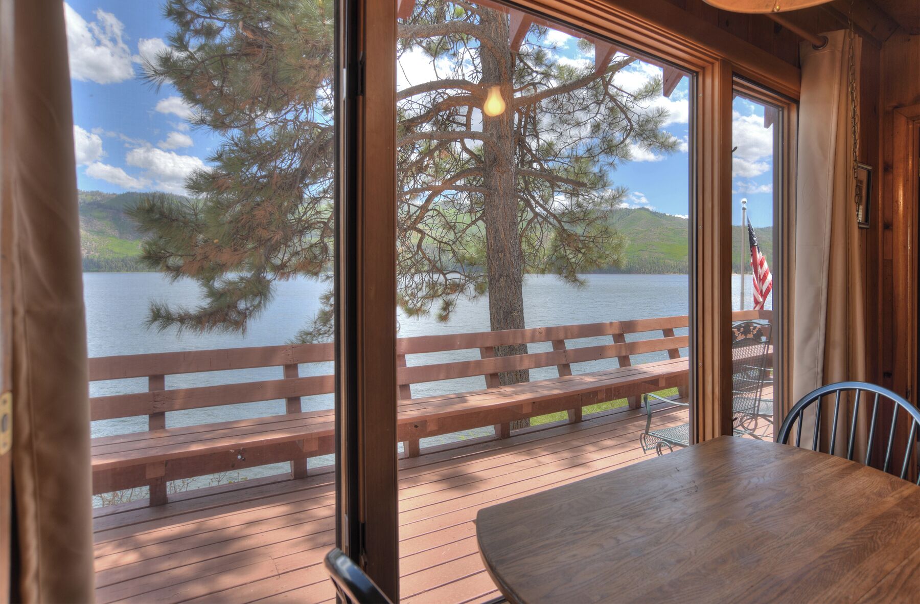 Um deck de madeira com mesa e cadeiras, com vista para um lago com uma árvore e um banco.