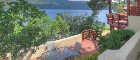 Une terrasse dotée d’une table et de chaises en verre, offrant une vue sur un lac, et entourée de verdure et d’une structure en bois.