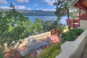 Eine Terrasse mit Glastisch und Stühlen mit Blick auf einen See, umgeben von Grünpflanzen und einer Holzhütte.