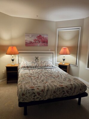 A bedroom with a bed, two bedside lamps, a nightstand, a wall-mounted painting, and a window with blinds.