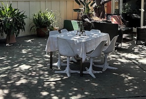 Ein runder Tisch mit weißer Tischdecke, umgeben von weißen Stühlen, auf einer Terrasses mit Topfpflanzen und einem Korbstuhl.