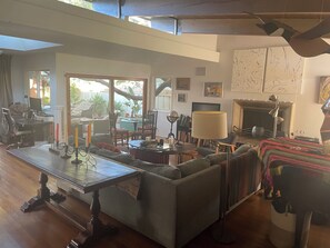 Ein großzügiger Wohnraum mit einer großen Couch, einem Esstisch und einem Kamin. Mehrere Stühle und ein kleiner Tisch mit Büchern und einer Lampe sind ebenfalls vorhanden.