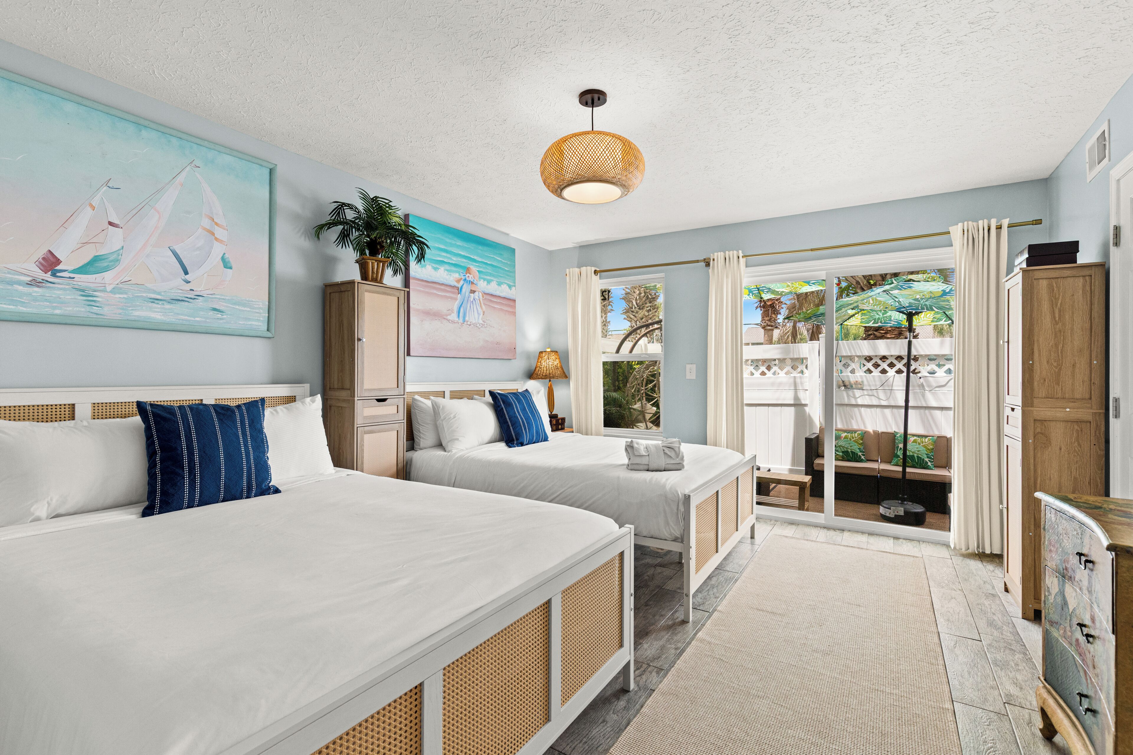 A hotel room with two beds, a large painting, a plant, and a view of the outdoors.
