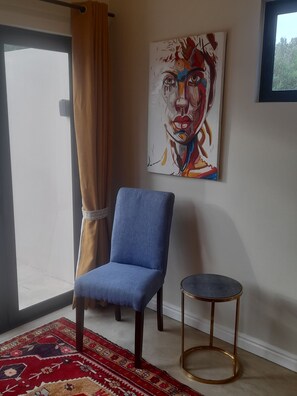 Habitació amb una cadira blava, una taula rodona petita, un quadre de paret colorit, una catifa amb dibuixos i una finestra amb cortines.