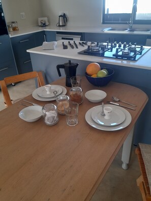 Uma mesa de jantar posta com pratos, copos e talheres, uma cafeteira e uma fruteira.