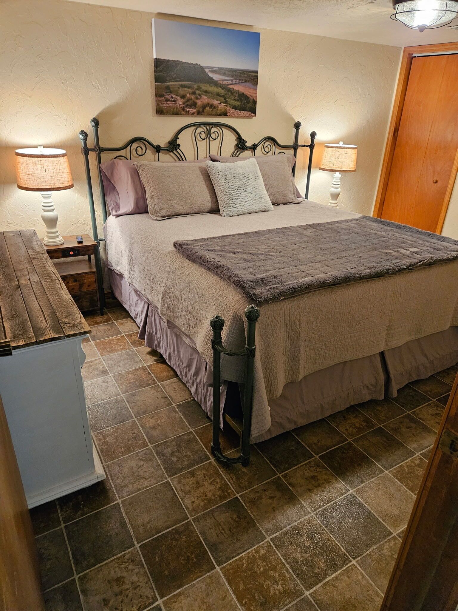 A bedroom with a bed, two bedside lamps, a wooden nightstand, a wooden dresser, and a door.