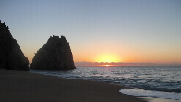Ein Strand mit Sonnenuntergang, Felsformationen und ruhiger See.