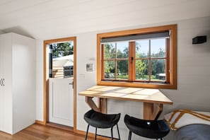 A compact kitchen with a wooden table and two black chairs, a white door, and a window with a view of trees. - Eldorado Caravan Park (Eldorado)