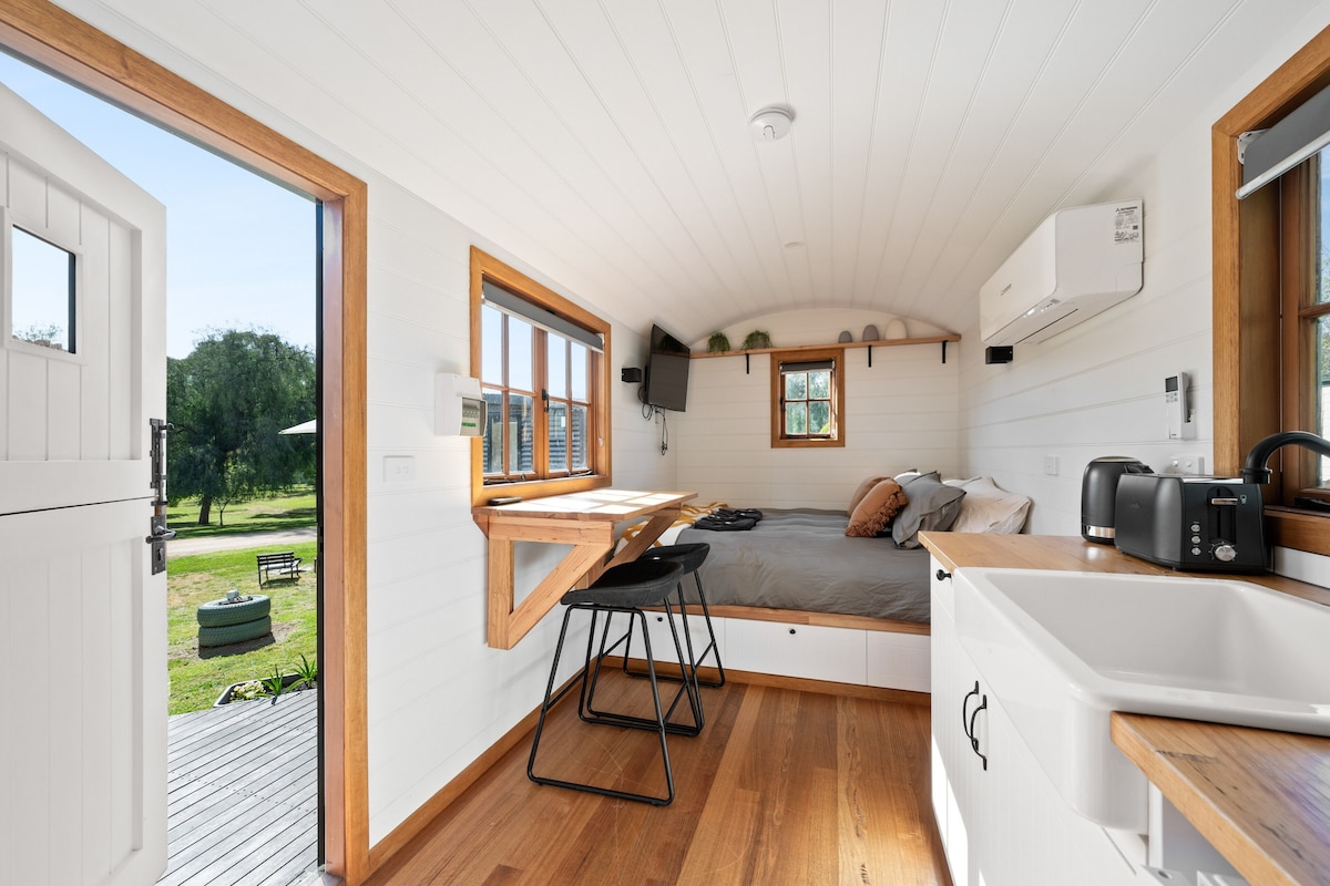 Uma cozinha compacta e moderna, com bancada de madeira, um pequeno espaço de refeições e uma cama com vista para o exterior.