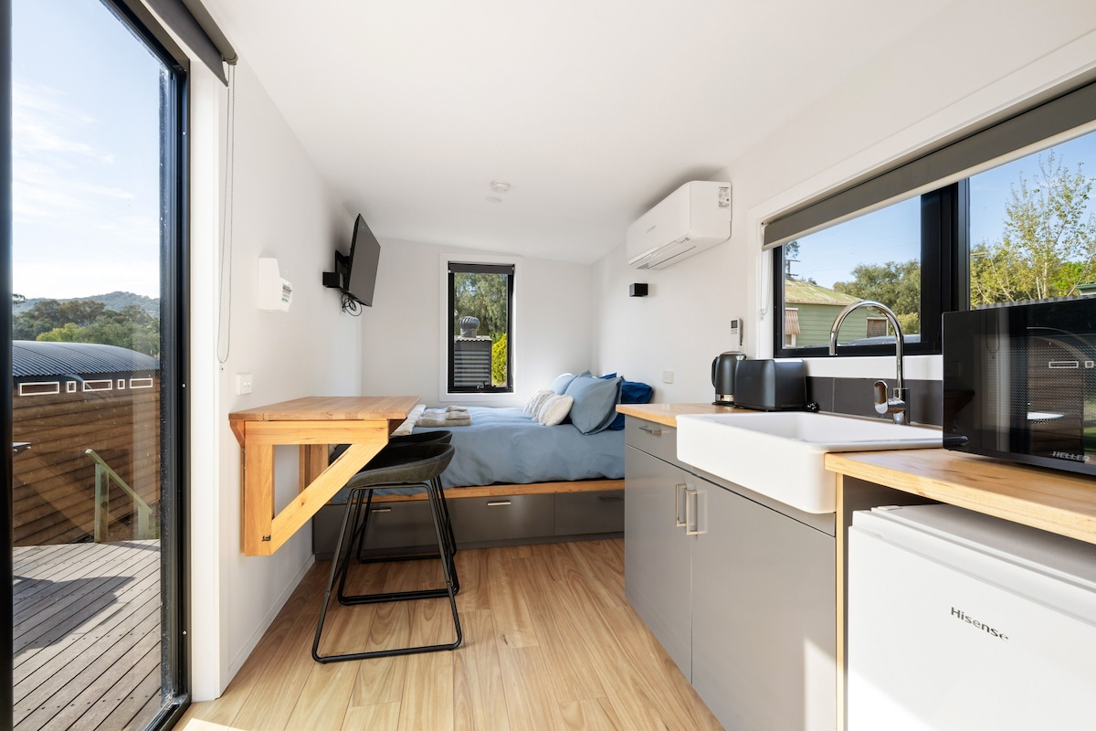 Uma cozinha compacta com uma mesa de madeira, uma cama com roupa de cama azul e vista para o exterior através de uma janela.