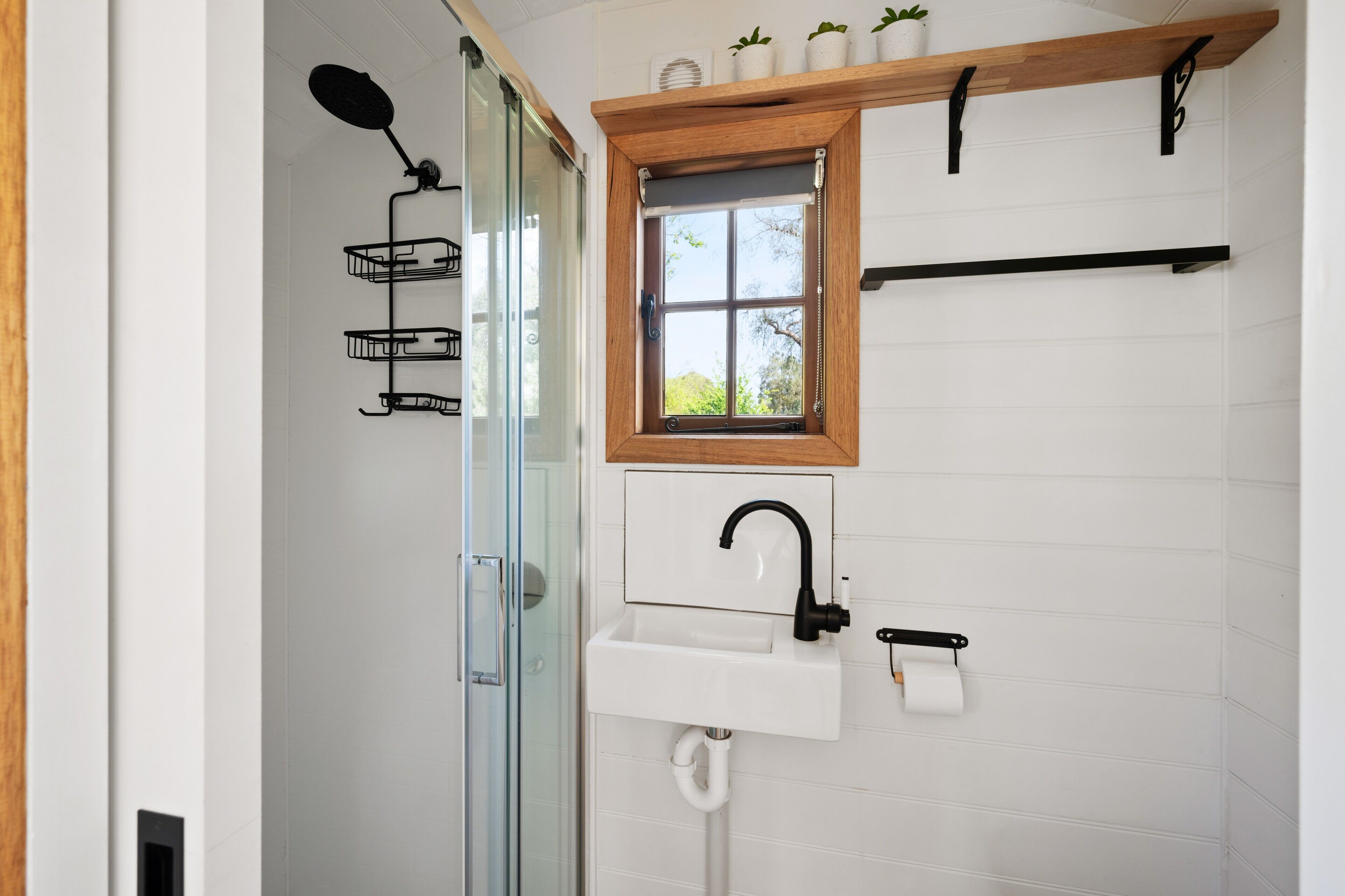 A bathroom with a glass shower enclosure, a white sink with a black faucet, and a small window above the sink.