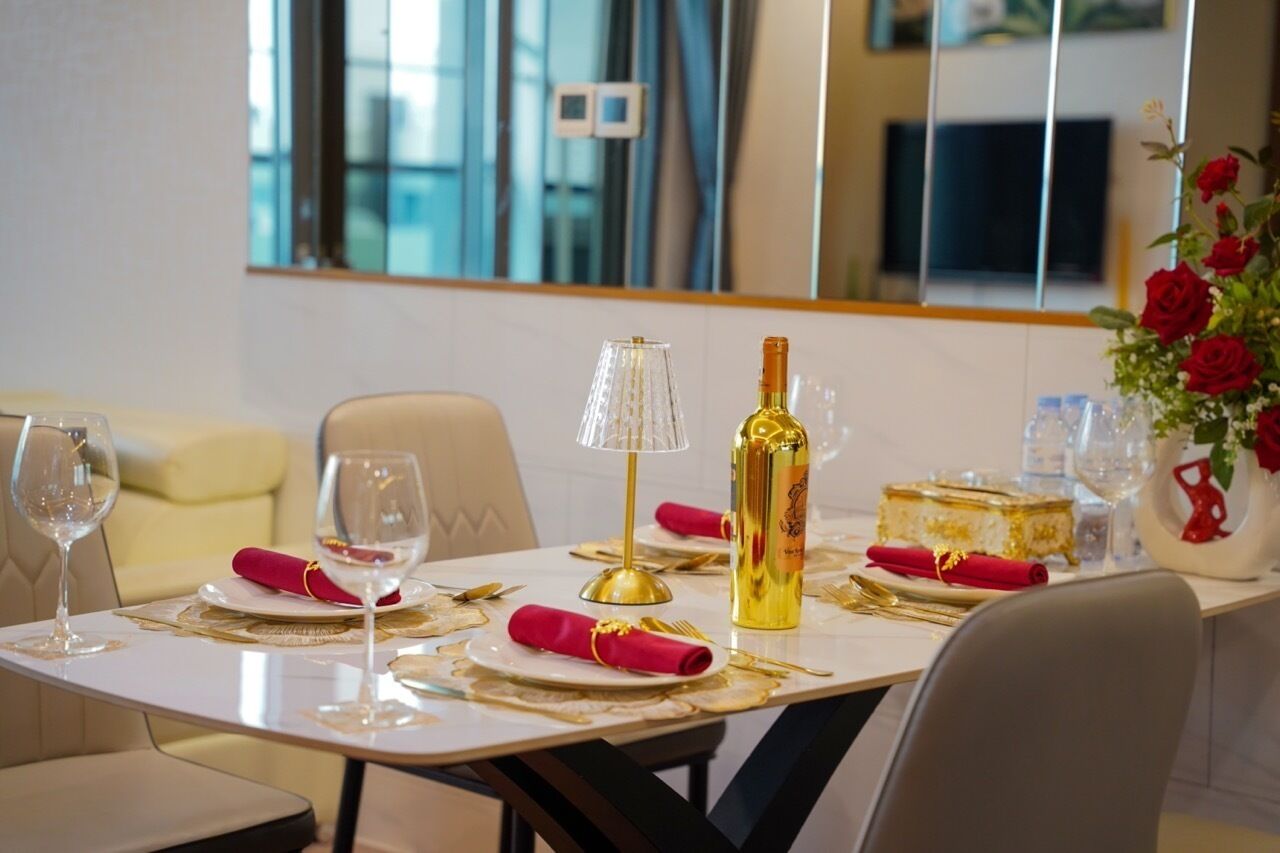 A dining table set with red napkins, gold cutlery, a bottle, and a cake, with a mirror and a vase of red roses in the background.
