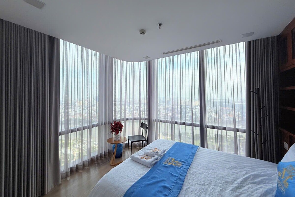 A hotel room with a large bed, a chair, a small table with a vase, and a view of the city through floor-to-ceiling windows.