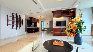 A modern living room with a sofa, a coffee table, and a TV. There is a kitchen area with cabinets and a dining table. - Landmark 81 Serviced Apartment - Mariana (Ho Chi Minh City)