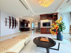 A modern living room with a sofa, a coffee table, and a TV. There is a kitchen area with cabinets and a dining table. - Landmark 81 Serviced Apartment - Mariana (Ho Chi Minh City)