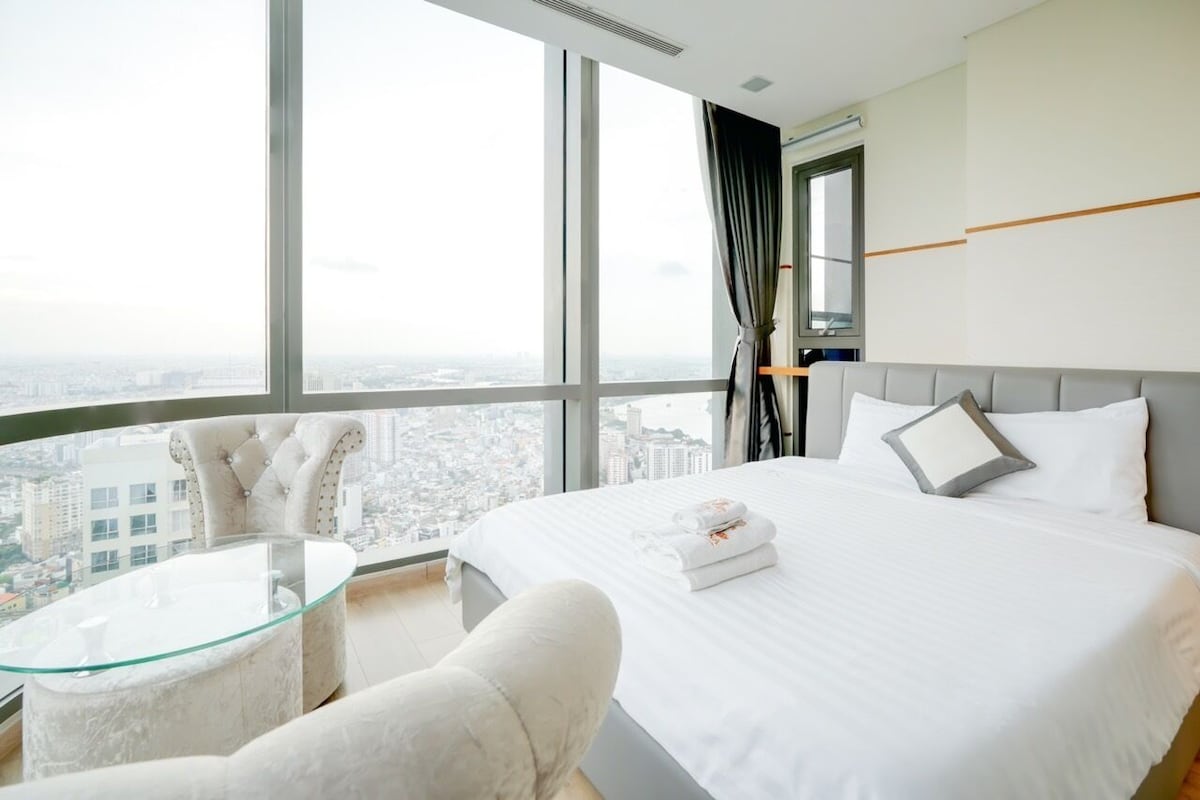 A hotel room with a large bed, a glass table, a chair, and a city view.