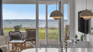 Habitación con un amplio ventanal corredera que da a una terraza con vista a un lago y colinas. Cuenta con una zona de comedor con mesa y sillas, y un televisor montado en la pared.