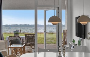 A room with a large sliding glass door leading to a deck with a view of a lake and hills. There is a dining area with a table and chairs, and a television mounted on the wall. - Amazing home in Ebeltoft with house sea view (Ebeltoft)