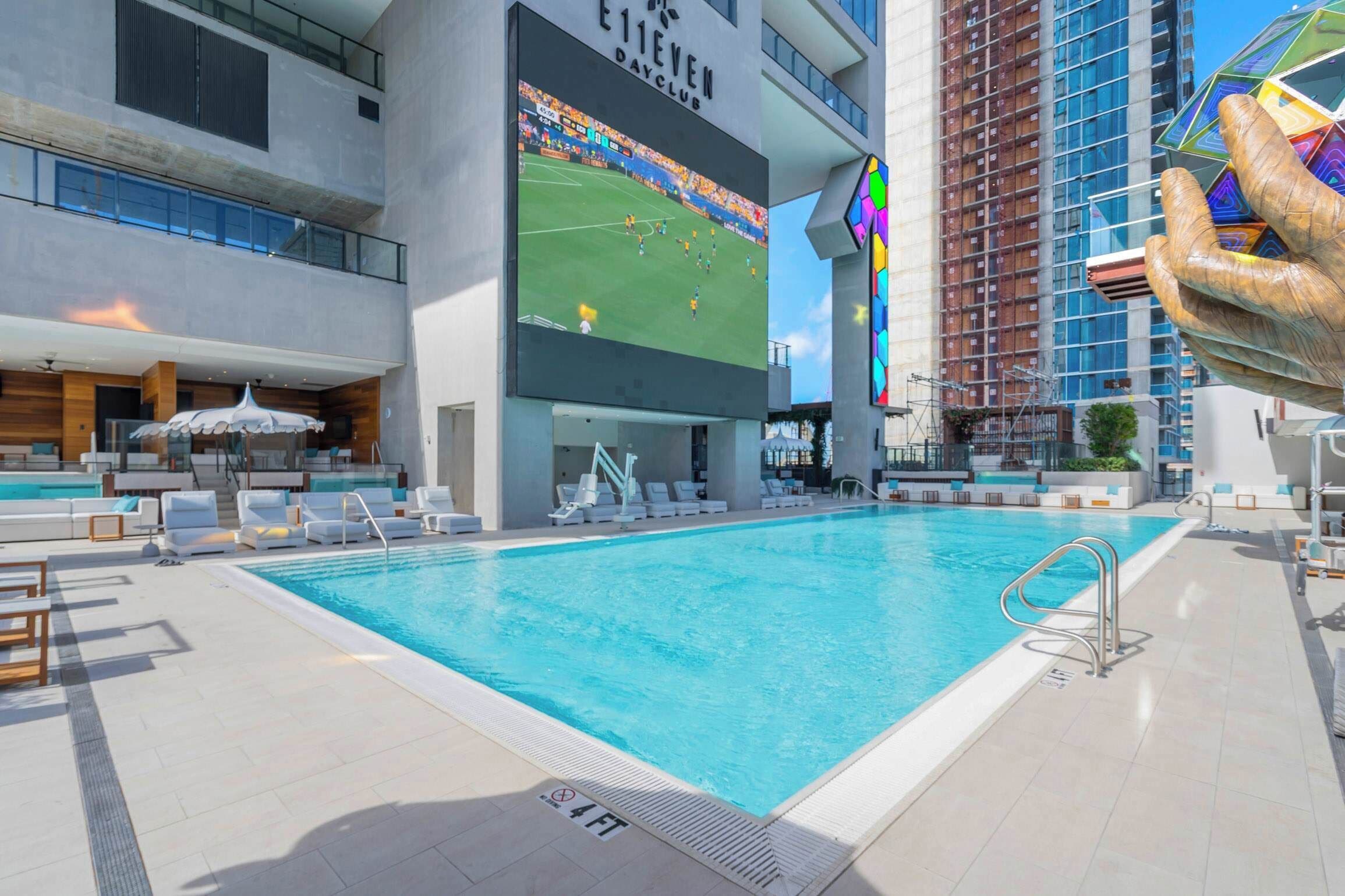 A swimming pool with lounge chairs, a large screen displaying a sports event, and a building with a sign reading 'E11EVEN DAYCLUB'.