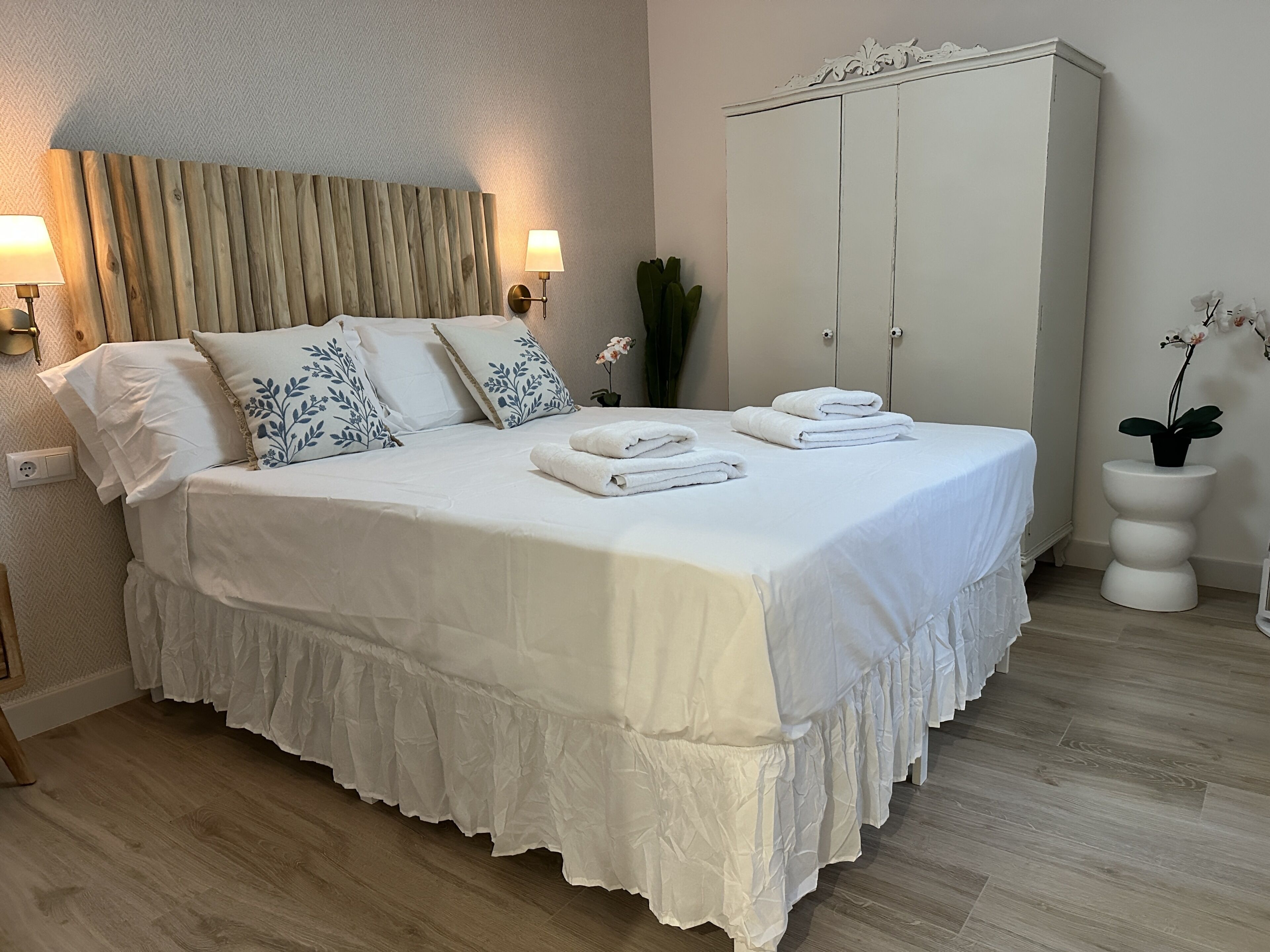 A neatly made bed with white linens and blue floral pillows, a bedside lamp, a white wardrobe, and a potted orchid.