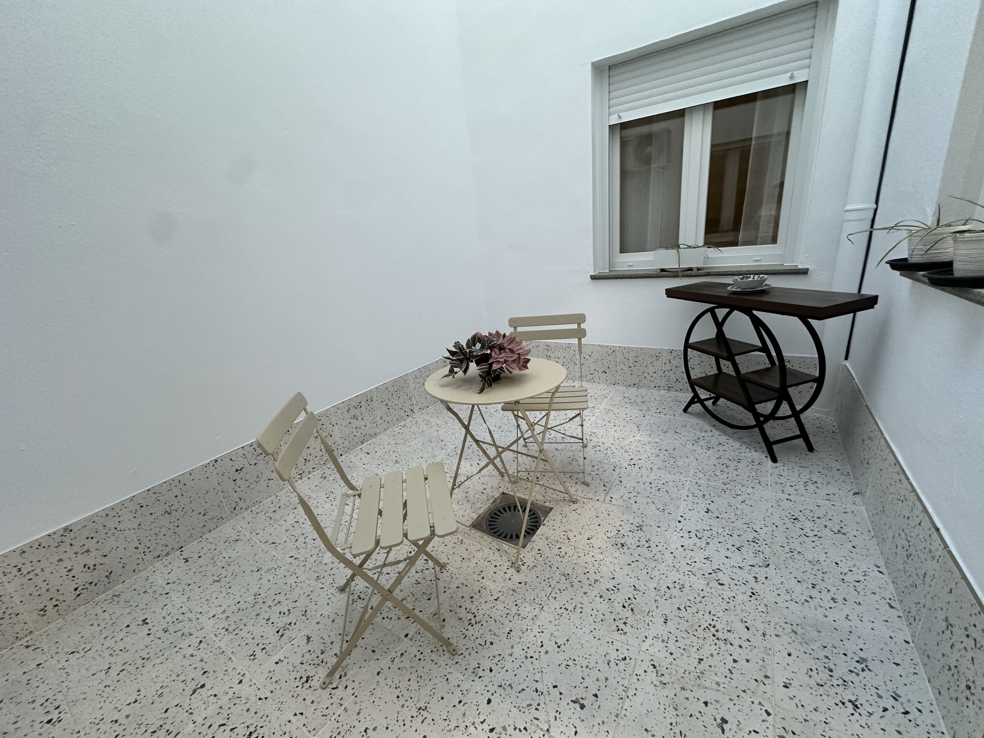 A small patio area with a round table, two chairs, a potted plant, and a window with blinds.