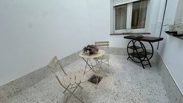 A small patio area with a round table, two chairs, a potted plant, and a window with blinds.