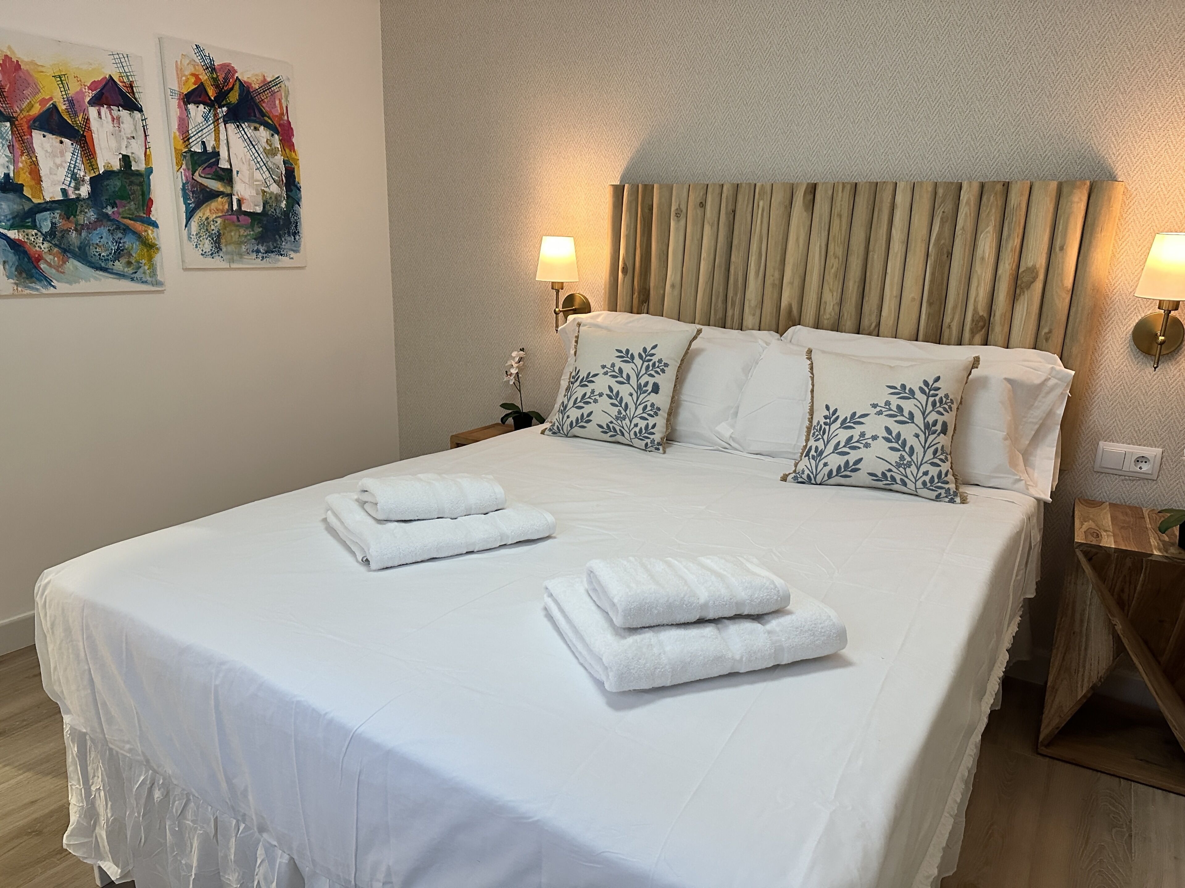 Una cama bien hecha con sábanas blancas y toallas dobladas, un cabecero de madera y dos lámparas fijadas a la pared.