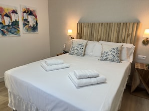 Una cama bien hecha con sábanas blancas y toallas dobladas, un cabecero de madera y dos lámparas fijadas a la pared.