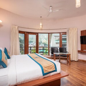 A hotel room with a large bed, a desk, and a chair, a ceiling fan, and large windows with curtains. - DLS Spring Valley Resort, Dharamshala (Dharamshala)