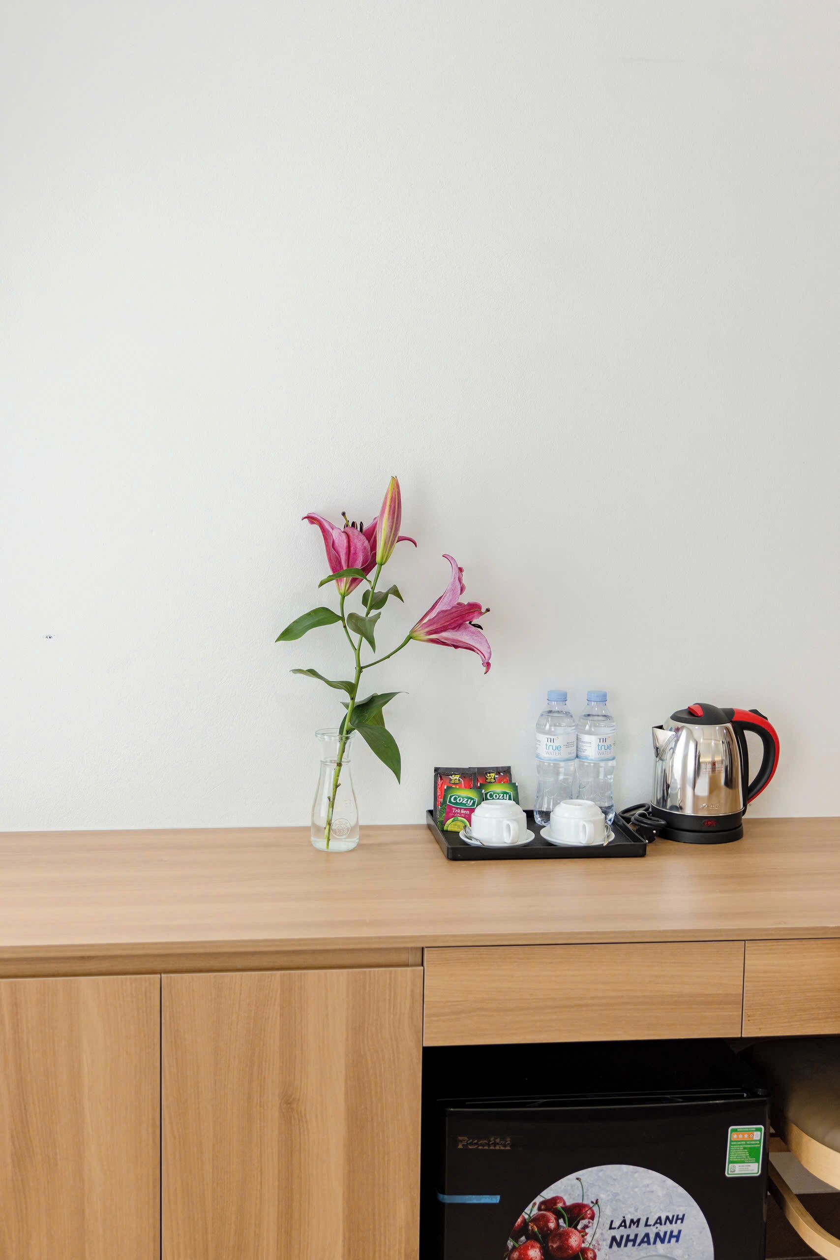 Una mesa de madera con un florero de flores rosas, una tetera, dos botellas de agua y una bandeja con tazas y una bolsa de aperitivos.