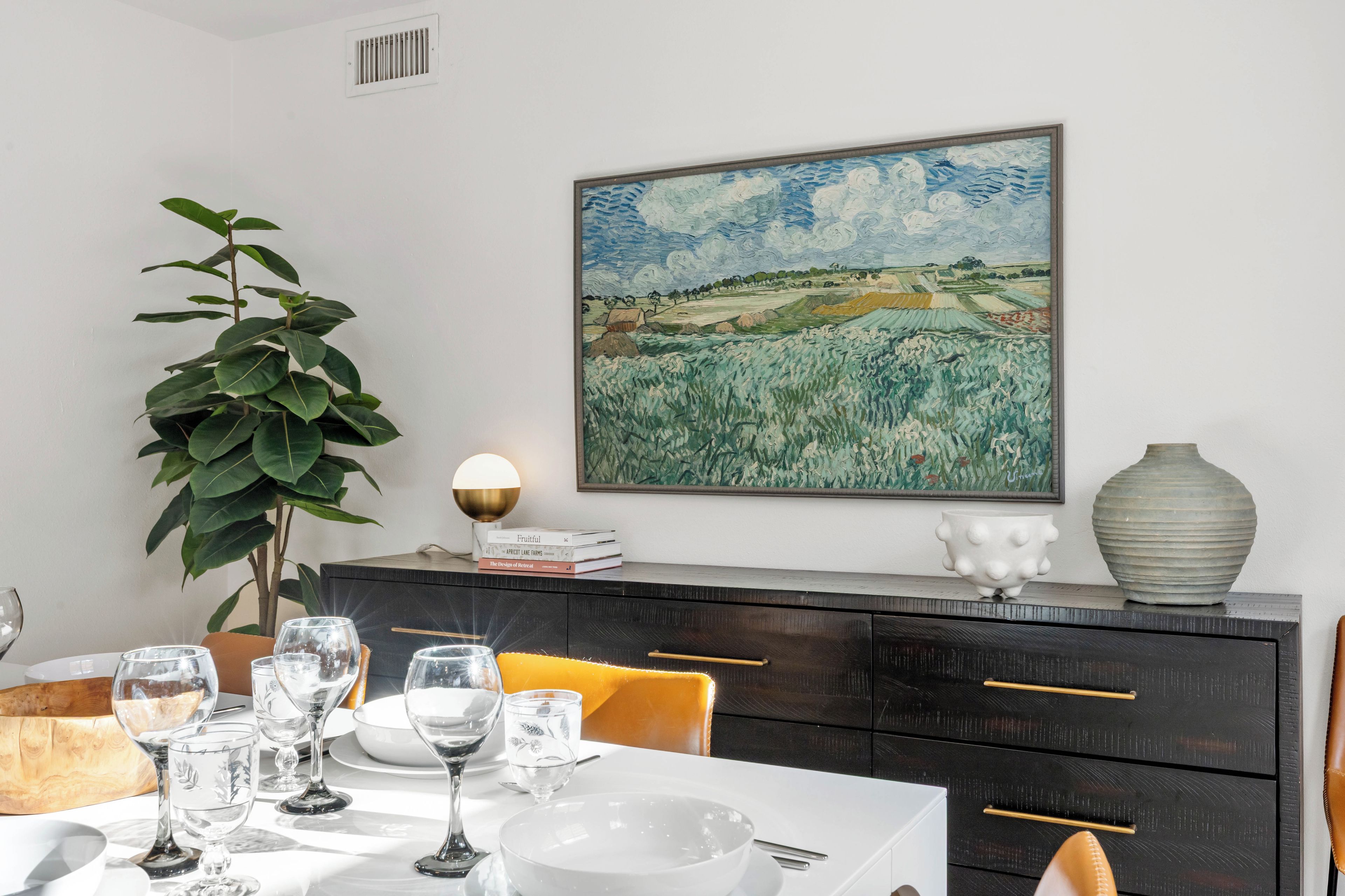 A dining area with a table set for four, a large painting on the wall, a potted plant, and a modern lamp.