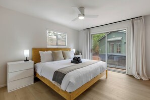 Ein Schlafzimmer mit einem hölzernen Bett, weißer Bettwäsche, einem Nachttisch, einem Deckenventilator und Blick auf ein grünes Haus durch große Fenster.