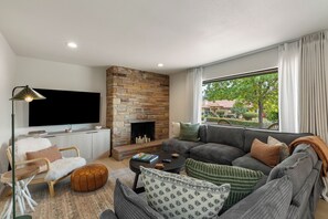 A modern living room with a stone fireplace, a large flat-screen TV, a sofa set, and a view of greenery outside. - West Sedona retreat with 4 bedrooms, hot tub, and Red Rock views. (Sedona)