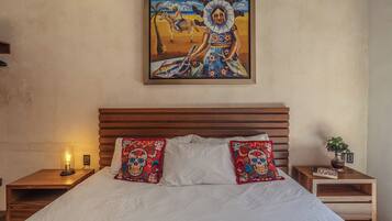 Un dormitorio con cabecera de madera, cama con ropa blanca, dos almohadones decorativos, una mesita de noche con lámpara y un cuadro en la pared.