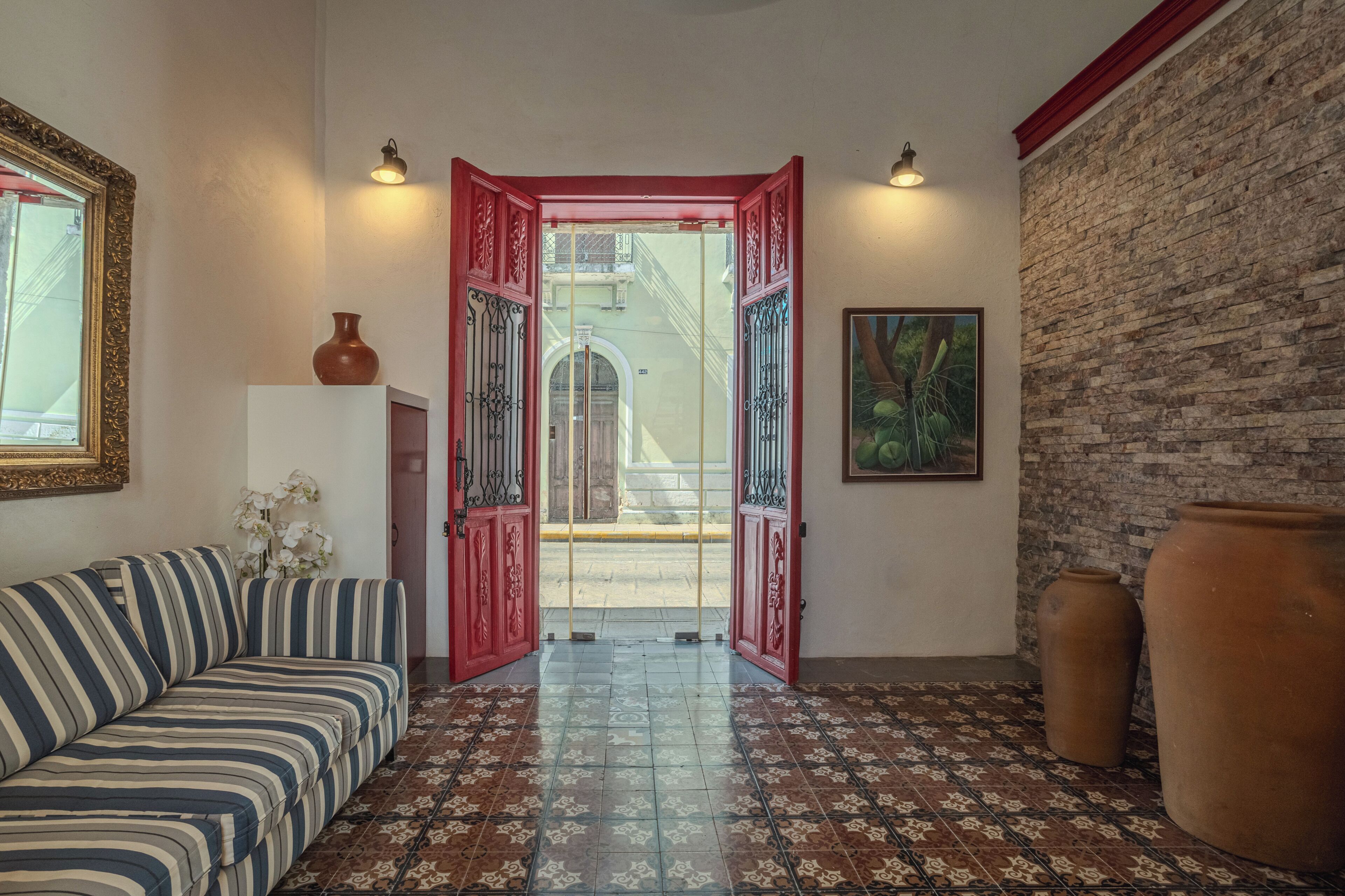 A room with a striped sofa, a tiled floor, a red door with decorative metalwork, a brick wall, and a framed picture.