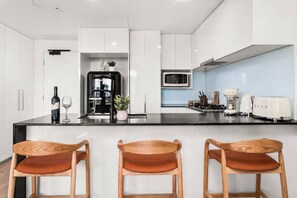 A modern kitchen with white cabinetry, a black countertop, and three wooden bar stools. - Luxury Meets Lifestyle (Fortitude Valley)
