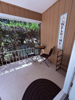 A small balcony with a table and two chairs, a welcome sign, and a view of trees.