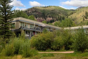 A two-story building with a balcony, surrounded by dense greenery and a mountain backdrop. - NEW! 1BR Mountain Retreat | Stunning Park & Mountain Views, Deck, Hot Tub + Steps to River (Telluride)