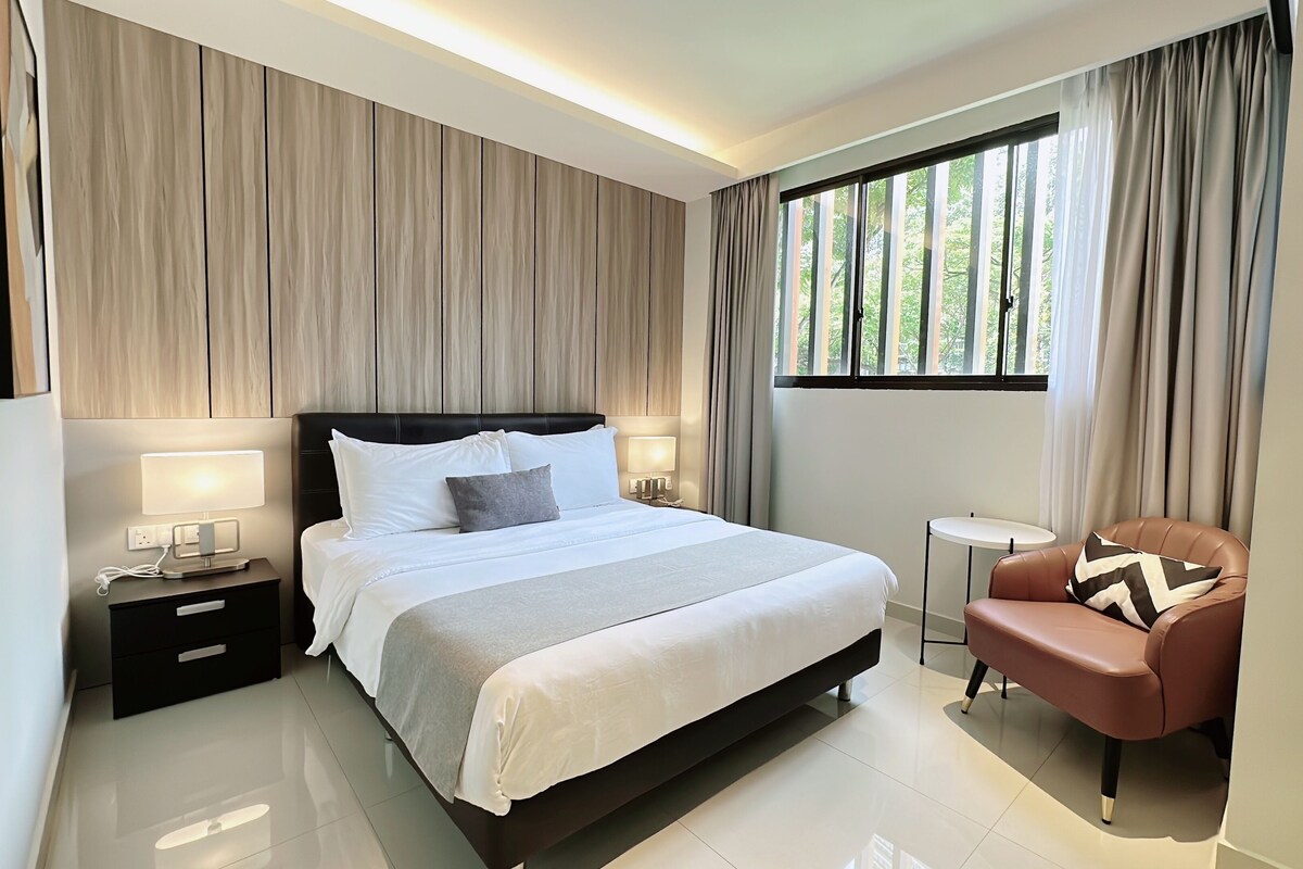 A modern hotel room with a large bed, bedside lamps, a small round table, and a leather armchair.