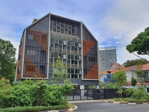A modern building with a glass facade and a geometric design, surrounded by greenery and other buildings.