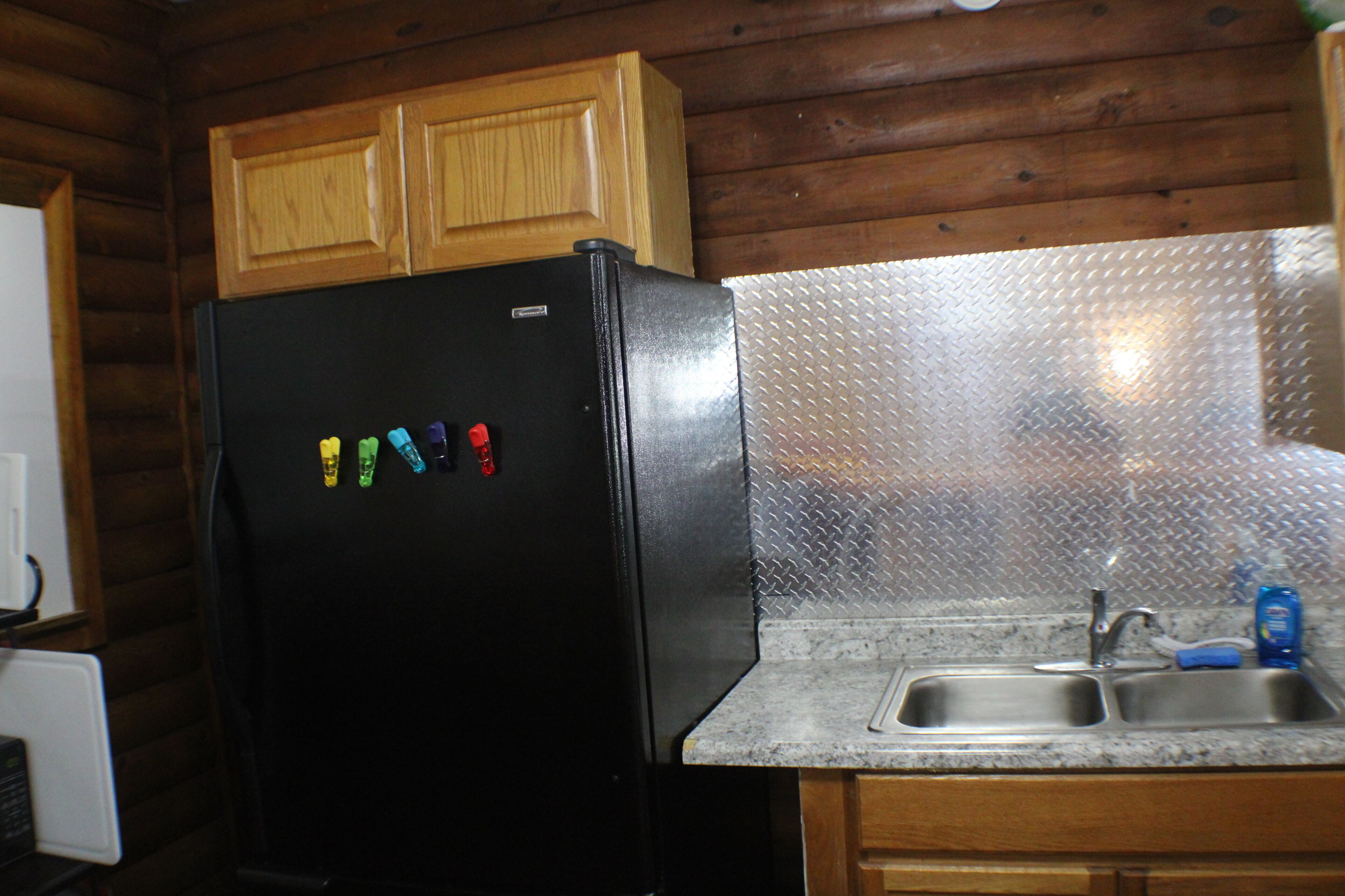 A kitchen with wooden cabinets, a black refrigerator, a granite countertop, and a stainless steel sink.