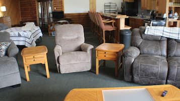 A cozy cabin interior with a bed, armchair, sofa, wooden side table, and a dining area with a table and chairs.