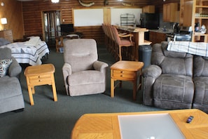 A cozy cabin interior with a bed, armchair, sofa, wooden side table, and a dining area with a table and chairs.